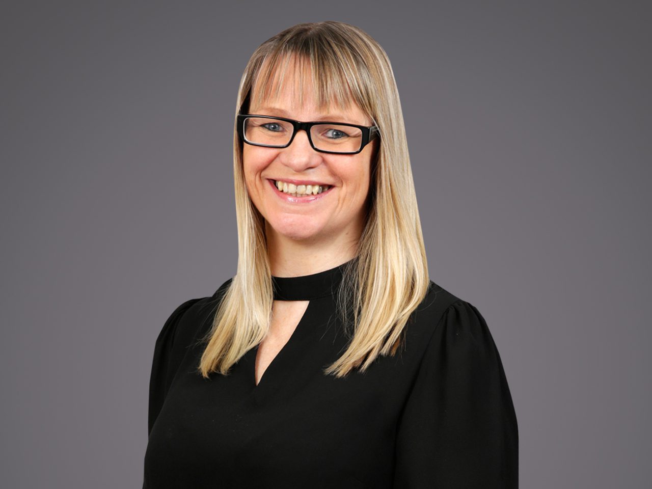 Smiling person with shoulder-length blond hair and black rectangular glasses, wearing a black top, against a neutral gray background.