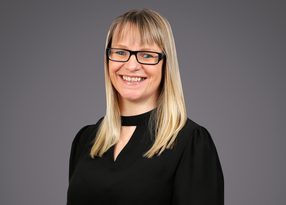 Smiling person with shoulder-length blond hair and black rectangular glasses, wearing a black top, against a neutral gray background.