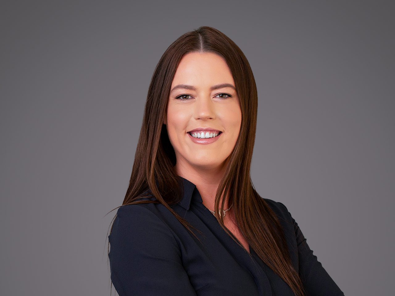 Smiling person with long brown hair in a dark blouse against a neutral gray studio backdrop.