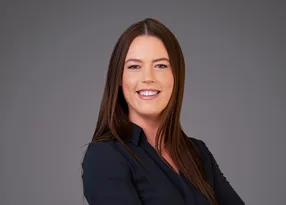 Portrait of a person with long brown hair, wearing a dark shirt, smiling against a neutral grey backdrop.