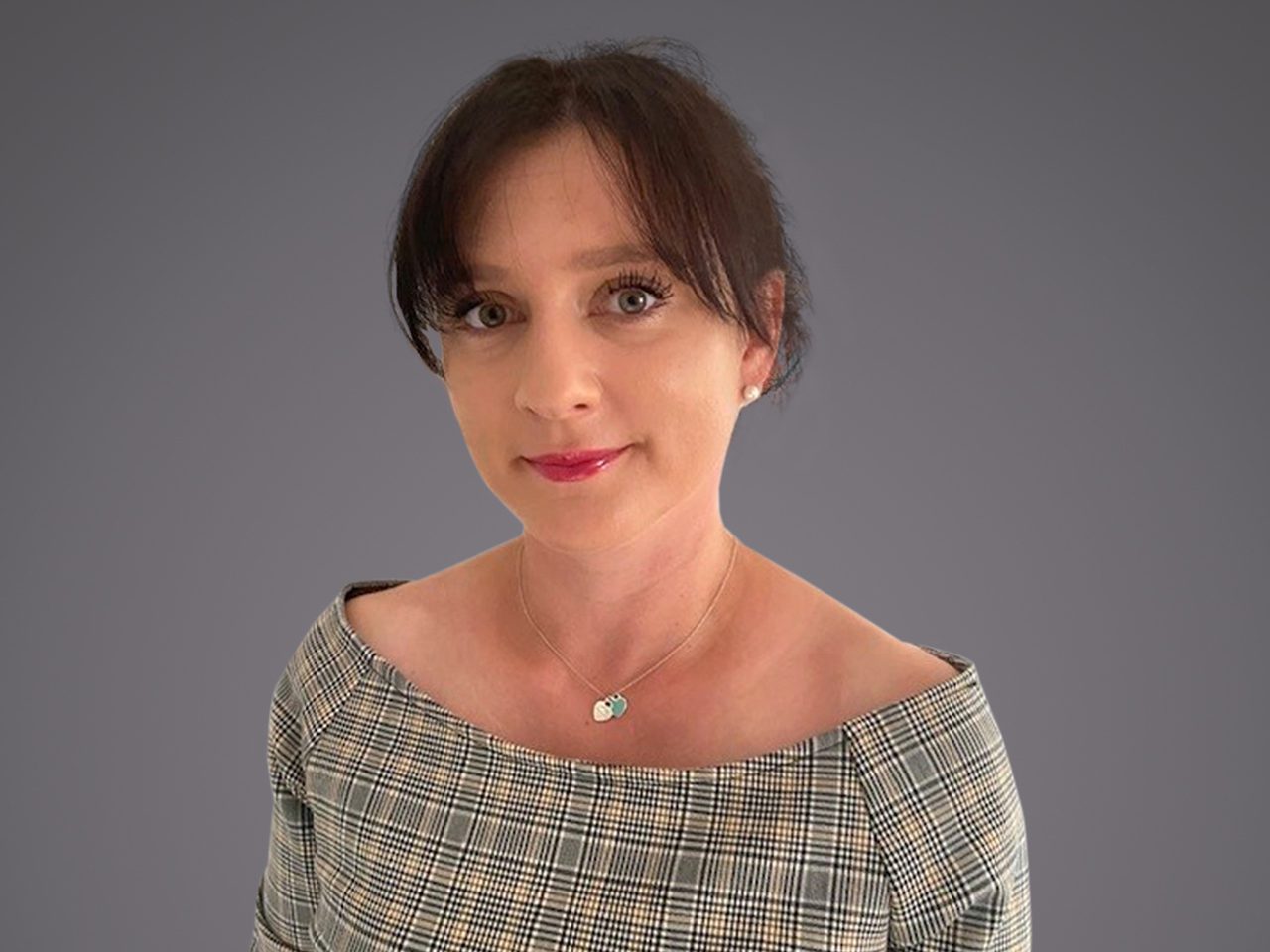 Portrait of a person with dark hair and light skin, wearing a plaid off-shoulder top and a small pendant necklace, smiling softly against a grey background.
