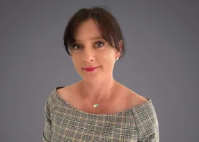 Portrait of a person with dark hair and light skin, wearing a plaid off-shoulder top and a small pendant necklace, smiling softly against a grey background.