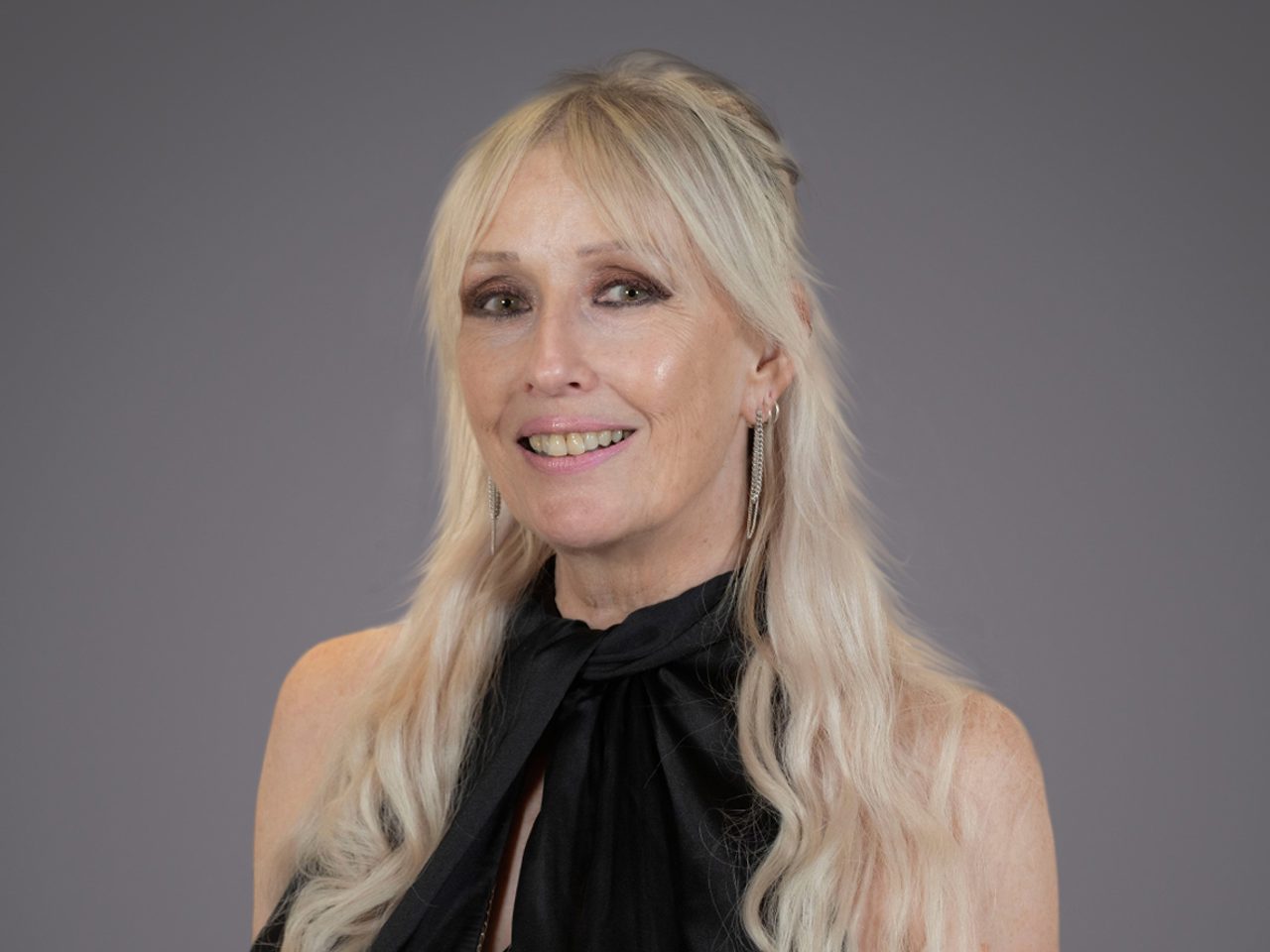 Portrait of a person with long blonde hair, wearing a black halter-neck top with a bow and silver drop earrings, smiling against a grey backdrop.