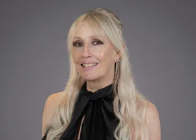 Portrait of a person with long blonde hair, wearing a black halter-neck top with a bow and silver drop earrings, smiling against a grey backdrop.