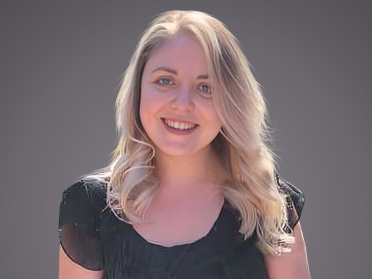 Une personne souriante avec des cheveux blonds ondulés porte un haut noir, sur un fond gris uni.