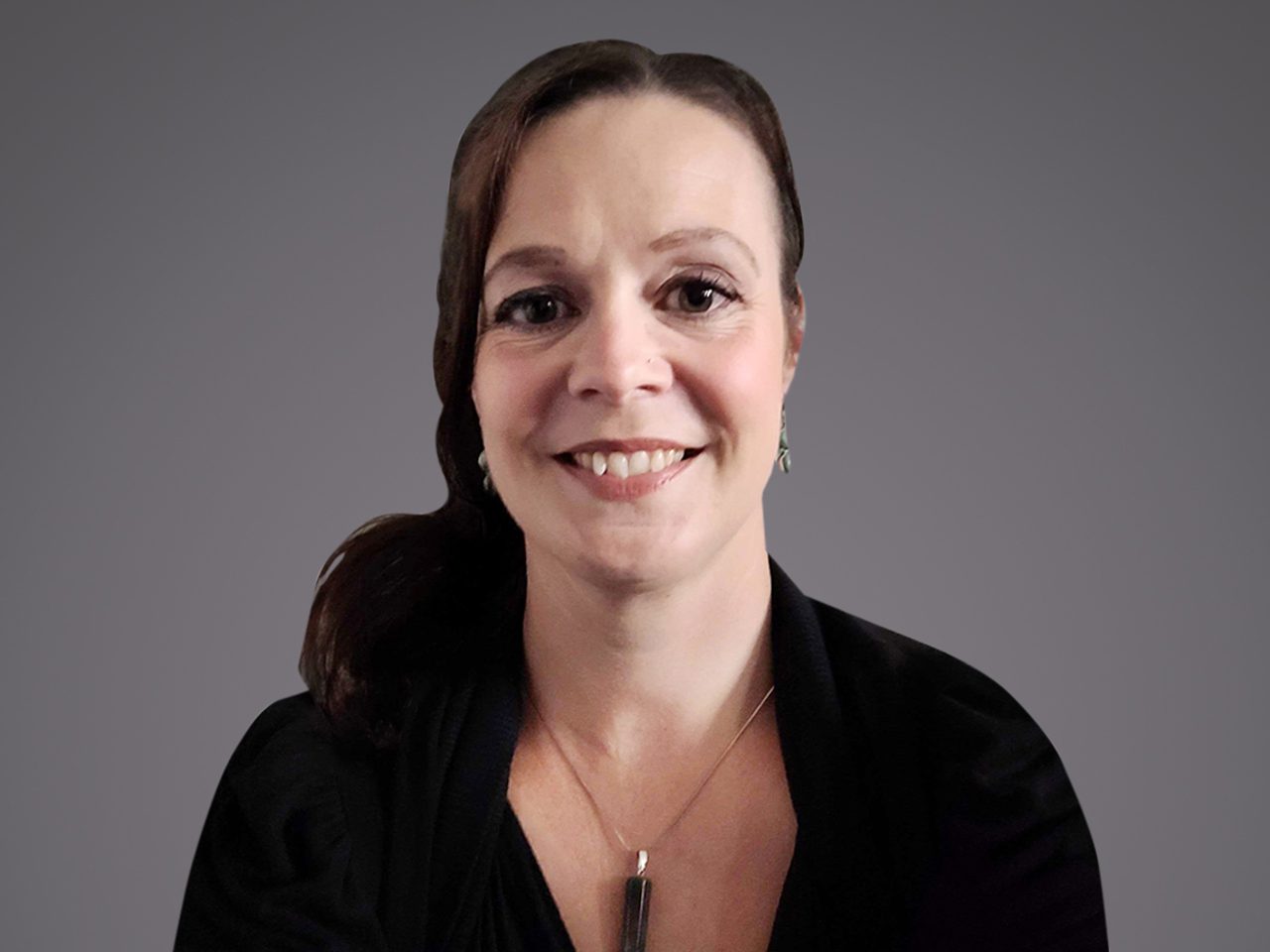 Person mit braunen Haaren, lächelnd, trägt schwarze Kleidung, silberne Ohrringe und eine lange silberne Kette mit rechteckigem Anhänger vor grauem Hintergrund.