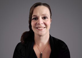 A person with dark brown hair tied back, wearing a black blazer, dangling earrings, and a pendant necklace, smiling against a gray background.