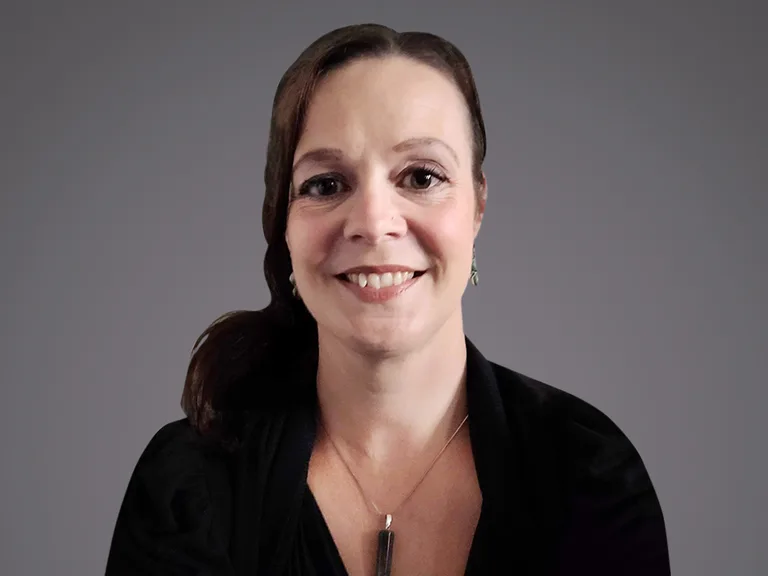 Ritratto di una persona sorridente con capelli castani raccolti, indossa top nero, orecchini e un pendente rettangolare, sfondo grigio sfumato.