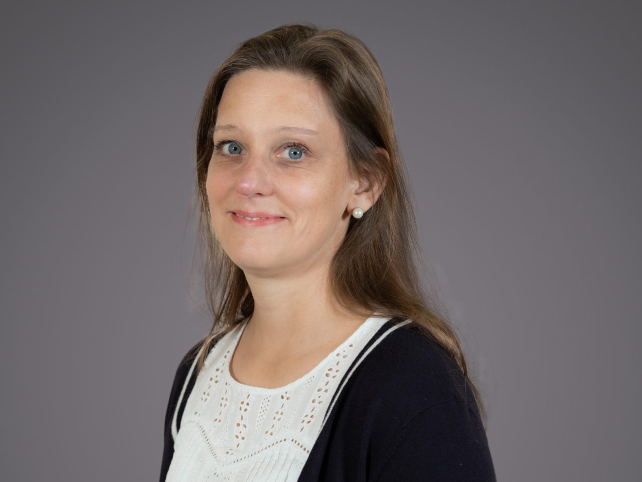 Portrait of a person with light brown hair and blue eyes, wearing a white lace blouse and dark cardigan with pearl earrings, smiling softly against a gray backdrop.
