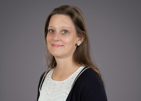 Portrait of a person with light brown hair and blue eyes, wearing a white lace blouse and dark cardigan with pearl earrings, smiling softly against a gray backdrop.