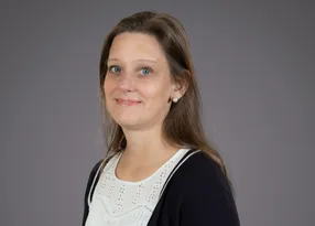 Person with shoulder-length brown hair and blue eyes, wearing a white lace blouse and dark cardigan, smiling against a grey background.