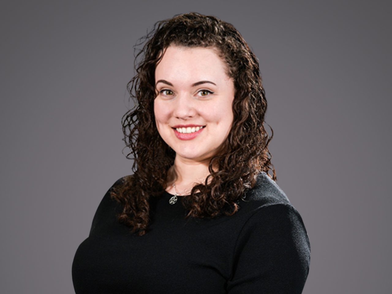 Portrait of a person with dark, curly hair wearing a black top, smiling at the camera against a grey background.