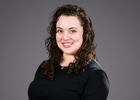 Portrait of a person with dark, curly hair wearing a black top, smiling at the camera against a grey background.