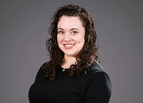 Portrait of a person with dark, curly hair wearing a black top, smiling at the camera against a grey background.