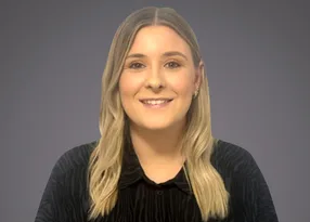 Smiling person with shoulder-length blond hair wearing a black top, against a neutral grey backdrop.