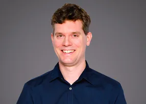 Smiling person with short brown hair wearing a navy shirt, posed against a neutral grey backdrop.
