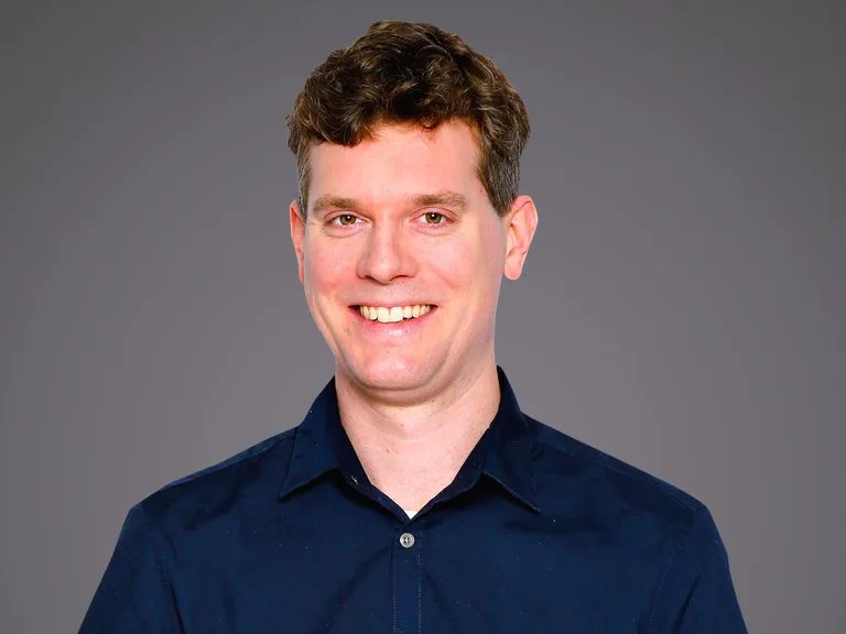 Portrait d'une personne souriante de face, cheveux bruns courts, portant une chemise bleu marine, sur fond gris.