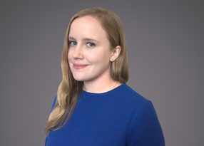 Portrait of a person with long blond hair wearing a blue blouse, facing slightly left with a soft smile, against a gray gradient backdrop.