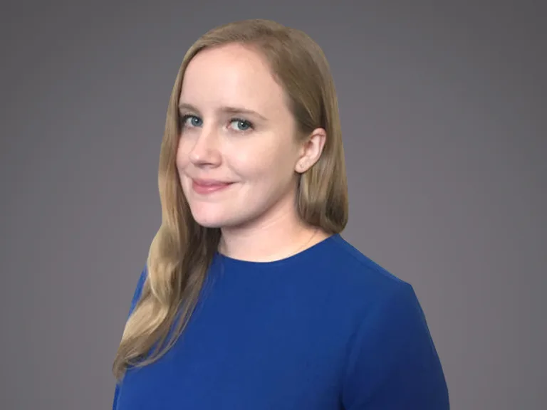 Portrait of a person with long blond hair wearing a blue blouse, facing slightly left with a soft smile, against a gray gradient backdrop.