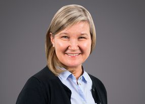 Portrait of a smiling person with light blonde hair, wearing a light blue button-up shirt and black cardigan, against a gray gradient backdrop.