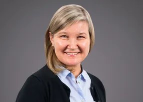 Portrait of a smiling person with blonde hair, wearing a light blue shirt and a dark cardigan, against a grey backdrop.