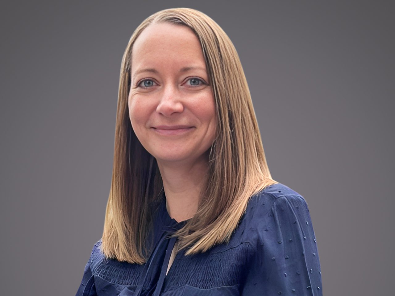 Portrait of a person with shoulder-length light brown hair and blue eyes, smiling softly, wearing a navy blouse with texture, against a gray gradient.