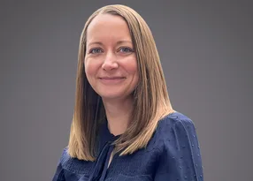 Head-and-shoulders portrait of a person with straight blonde hair, wearing a blue textured blouse with small dots, smiling against a grey gradient background.