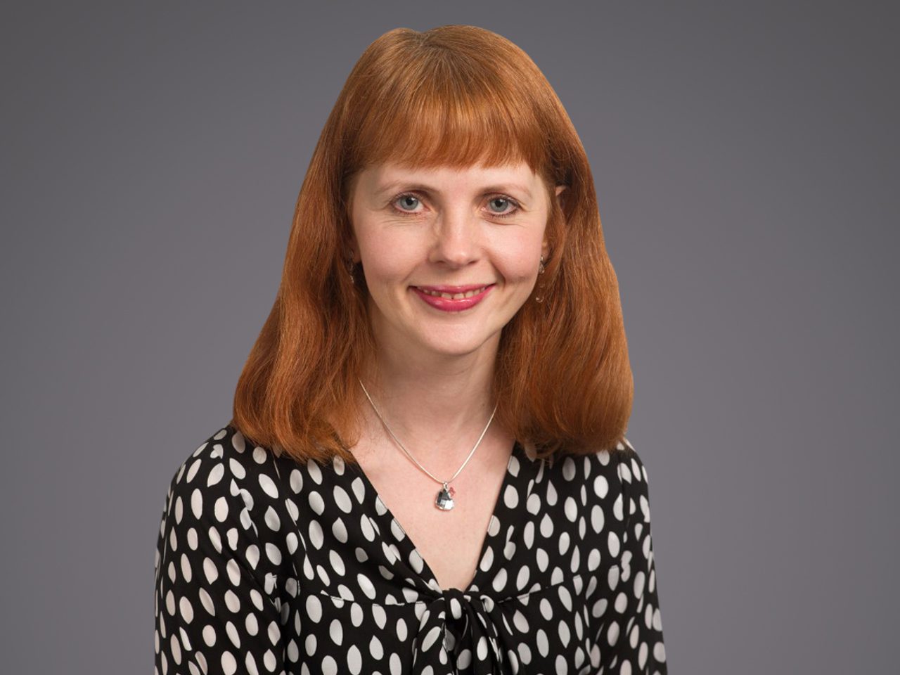 Portrait of a person with red shoulder-length hair, wearing a black-and-white polka-dot blouse and pendant necklace, smiling against a grey backdrop.