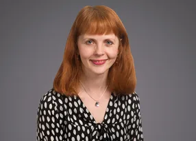 Portrait of a person with red shoulder-length hair, wearing a black-and-white polka-dot blouse and pendant necklace, smiling against a grey backdrop.