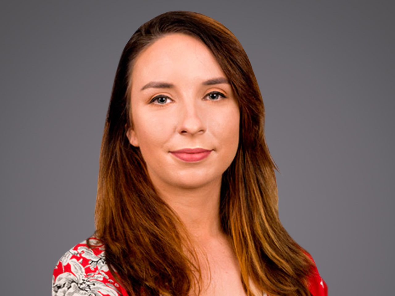 A person with shoulder-length brown hair wearing a red floral top, facing the camera with a calm expression against a gray background.
