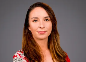 Portrait of a person with long brown hair wearing a red floral top, smiling softly, against a solid grey background.