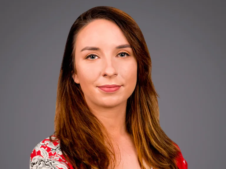 Ritratto frontale di una persona adulta con capelli castano chiaro lunghi, espressione neutra, indossa un top rosso con motivo floreale, sfondo grigio uniforme.