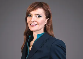 Portrait of a person with long auburn hair, wearing a navy blazer over a teal blouse, smiling against a grey studio background.