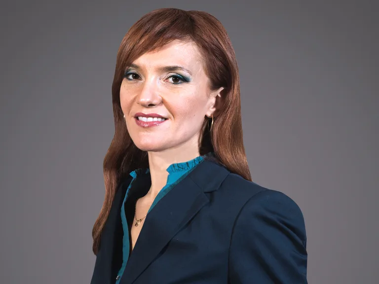 Portrait of a person with long auburn hair, wearing a navy blazer over a teal blouse, smiling against a grey studio background.