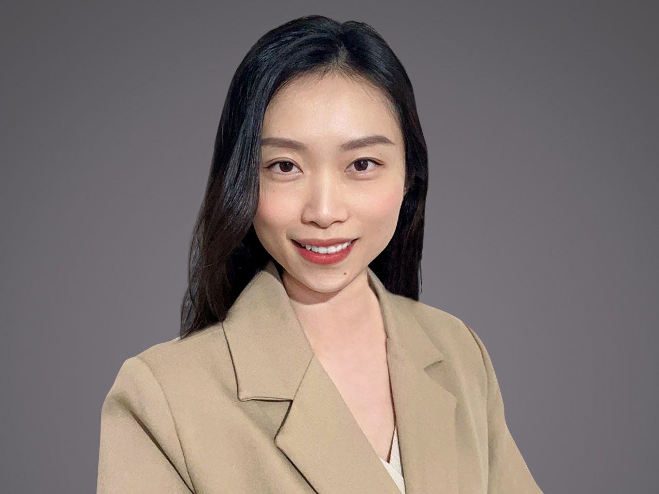 Portrait of a person with shoulder-length black hair, wearing a beige blazer over a white top, smiling at the camera against a grey background.