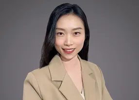 Portrait of a person with shoulder-length black hair, wearing a beige blazer over a white top, smiling at the camera against a grey background.