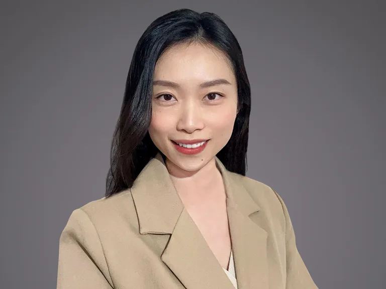 Portrait of a person with shoulder-length black hair, wearing a beige blazer over a white top, smiling at the camera against a grey background.
