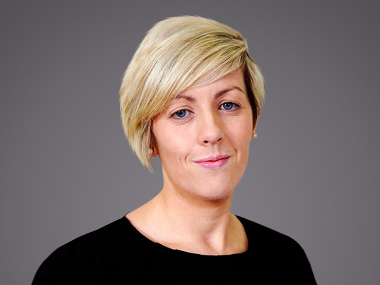 Head-and-shoulders portrait of a person with short blonde hair and blue eyes, wearing a black top, against a neutral grey background.