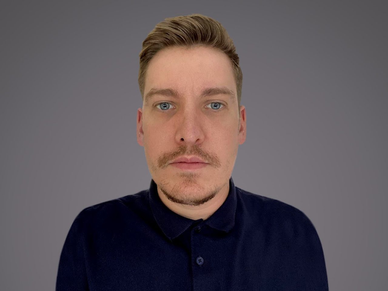 A person with short blond hair, blue eyes and light stubble, wearing a dark navy polo shirt, looking at the camera against a grey gradient background.