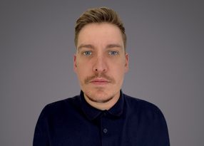 Close-up of a person with short blond hair and blue eyes, wearing a dark polo shirt, neutral expression, against a gray gradient background.