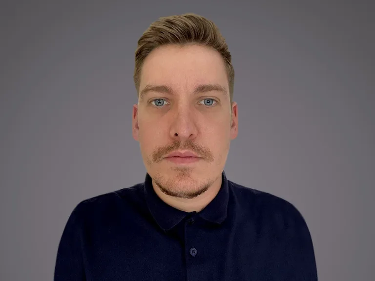 A person with short blond hair, blue eyes and light stubble, wearing a dark navy polo shirt, looking at the camera against a grey gradient background.