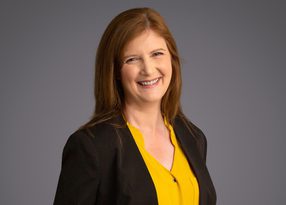 Smiling person with shoulder-length brown hair, wearing a yellow blouse and black blazer, against a neutral gray background.