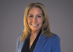 Portrait of a person with shoulder-length blonde hair, wearing a blue pinstripe blazer over a black blouse, smiling against a grey background.