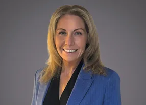 Portrait of a person with shoulder-length blonde hair, wearing a blue pinstripe blazer over a black blouse, smiling against a grey background.