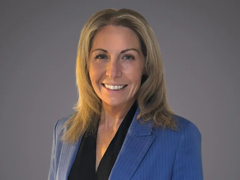Portrait of a person with shoulder-length blonde hair, wearing a blue pinstripe blazer over a black blouse, smiling against a grey background.