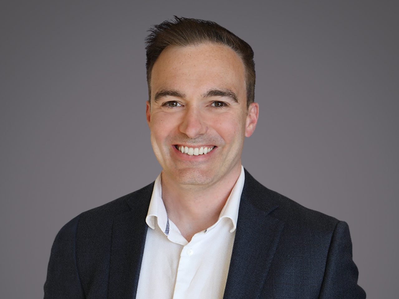 Smiling professional portrait of a person with short dark hair, wearing a dark blazer and white shirt, against a grey gradient background.