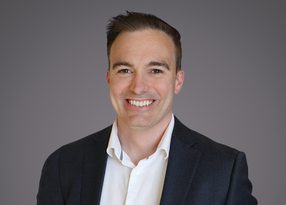 Smiling professional portrait of a person with short dark hair, wearing a dark blazer and white shirt, against a grey gradient background.