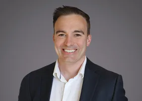 Smiling professional portrait of a person with short dark hair, wearing a dark blazer and white shirt, against a grey gradient background.