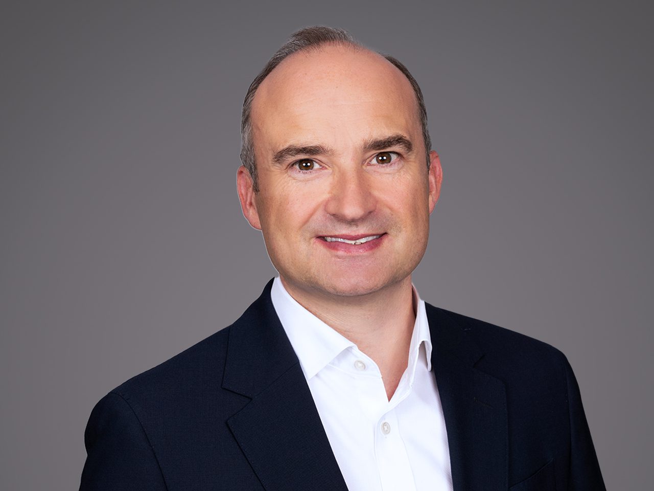 Portrait of a person with a friendly smile, wearing a dark blazer and white shirt, against a neutral gray background.
