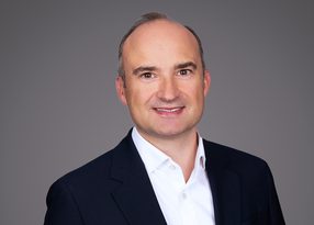 Head-and-shoulders portrait of a smiling person in a white shirt and dark blazer against a grey studio background.